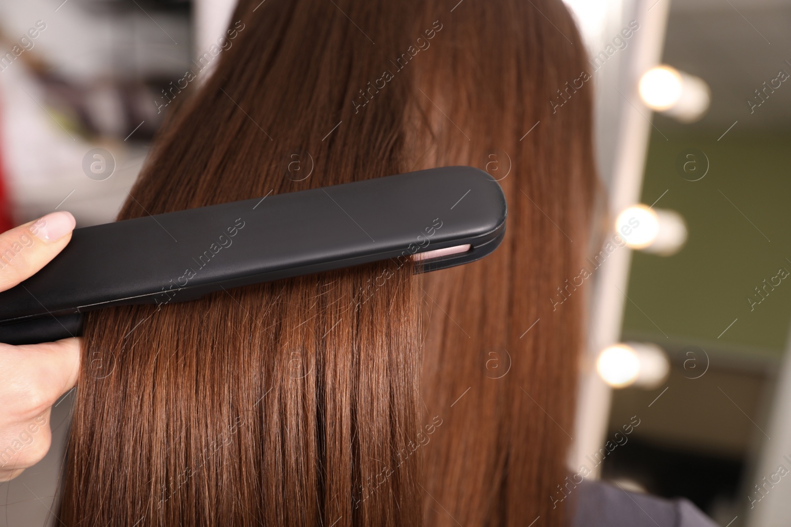 Photo of Stylist straightening woman's hair with flat iron in salon, closeup