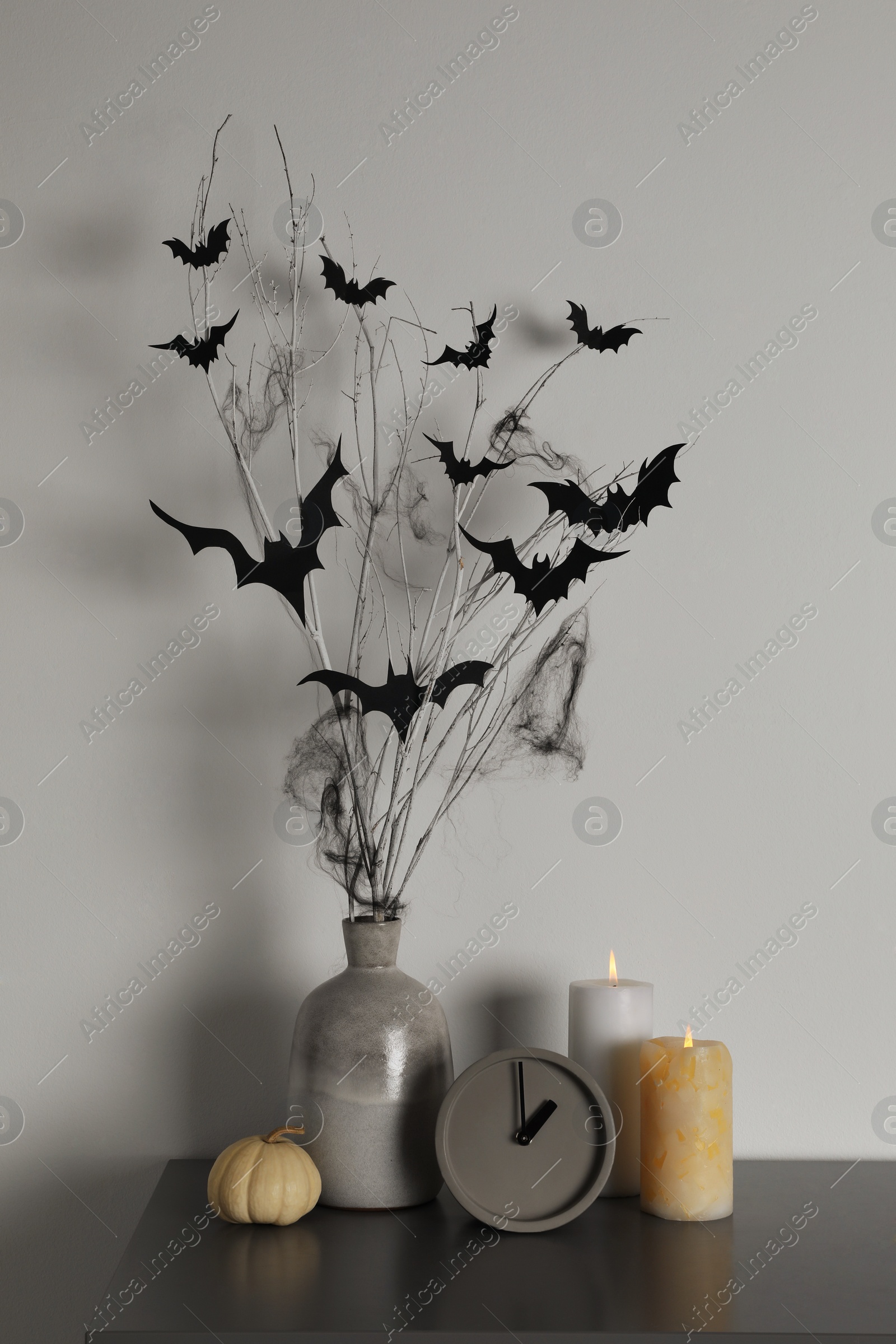 Photo of Alarm clock, burning candles and vase with paper bats  on table near white wall. Halloween decor