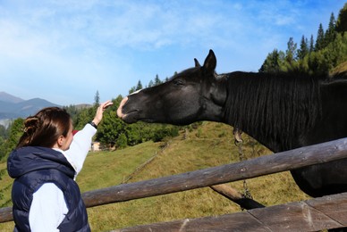 Photo of Woman stroking black horse near wooden fence in mountains. Beautiful pet