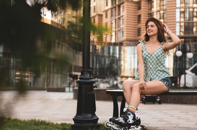 Beautiful young woman with roller skates sitting on bench outdoors, space for text