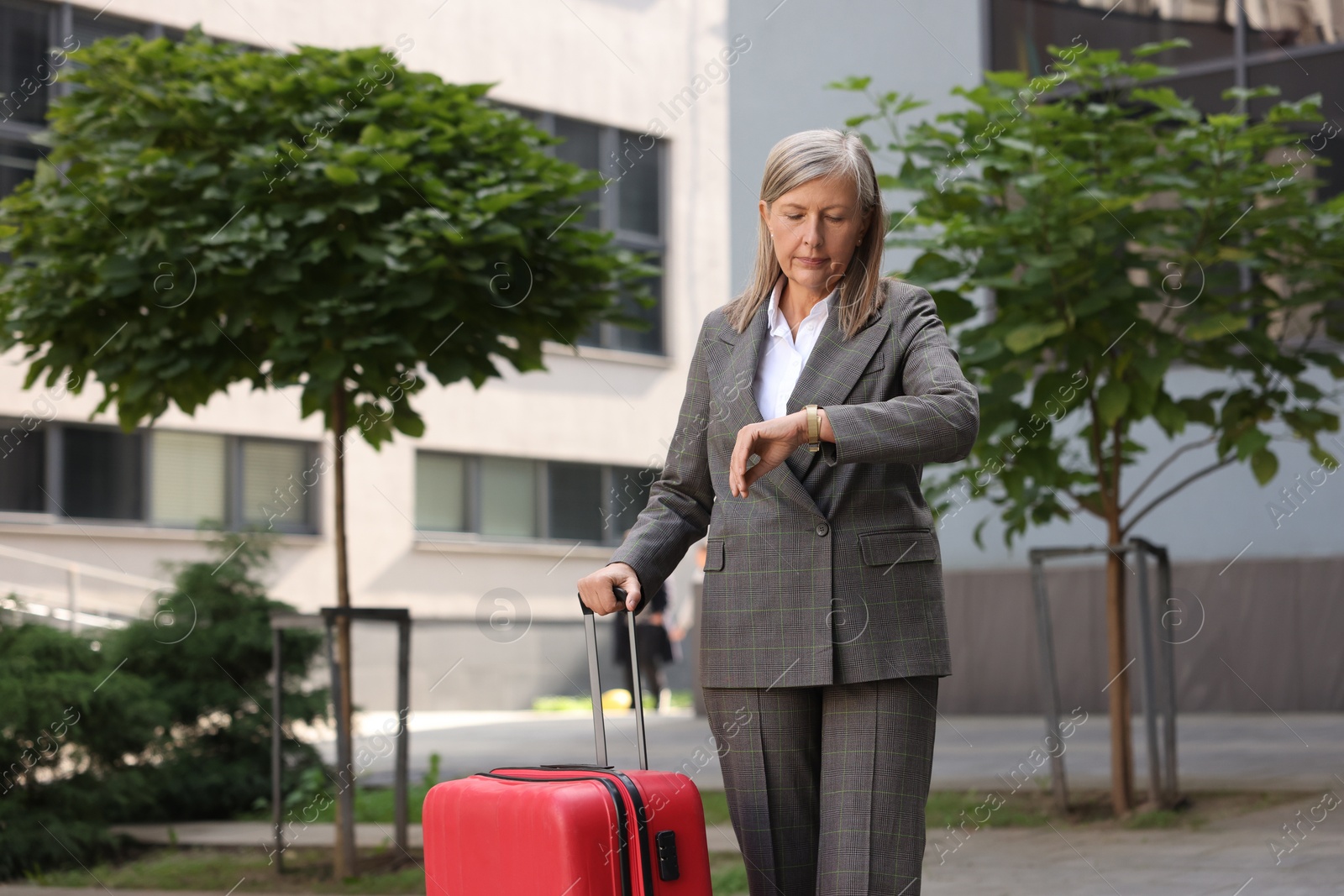 Photo of Being late. Worried senior businesswoman with red suitcase looking at watch outdoors