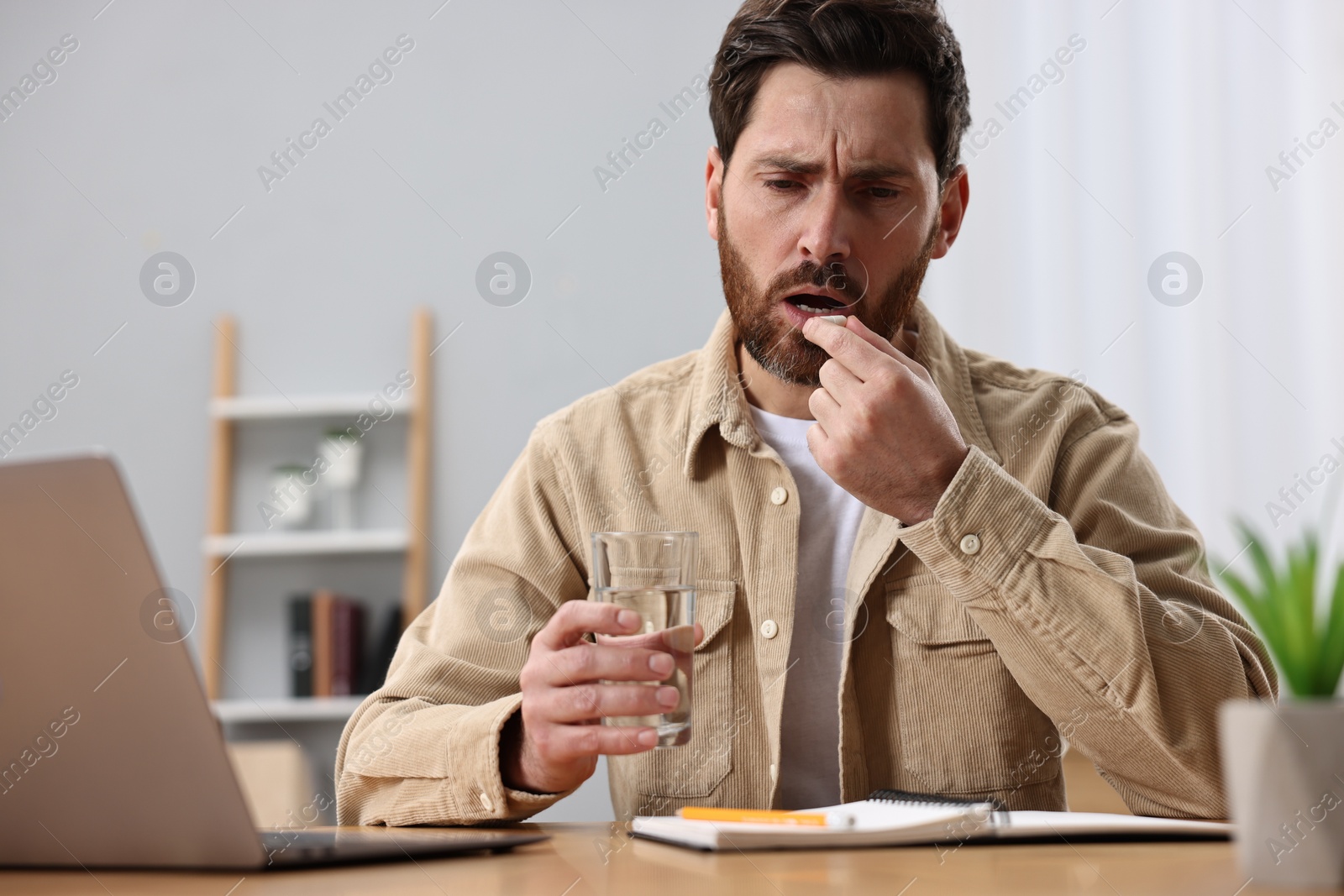 Photo of Worried man with glass of water taking pill from headache at workplace.