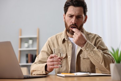 Photo of Worried man with glass of water taking pill from headache at workplace.