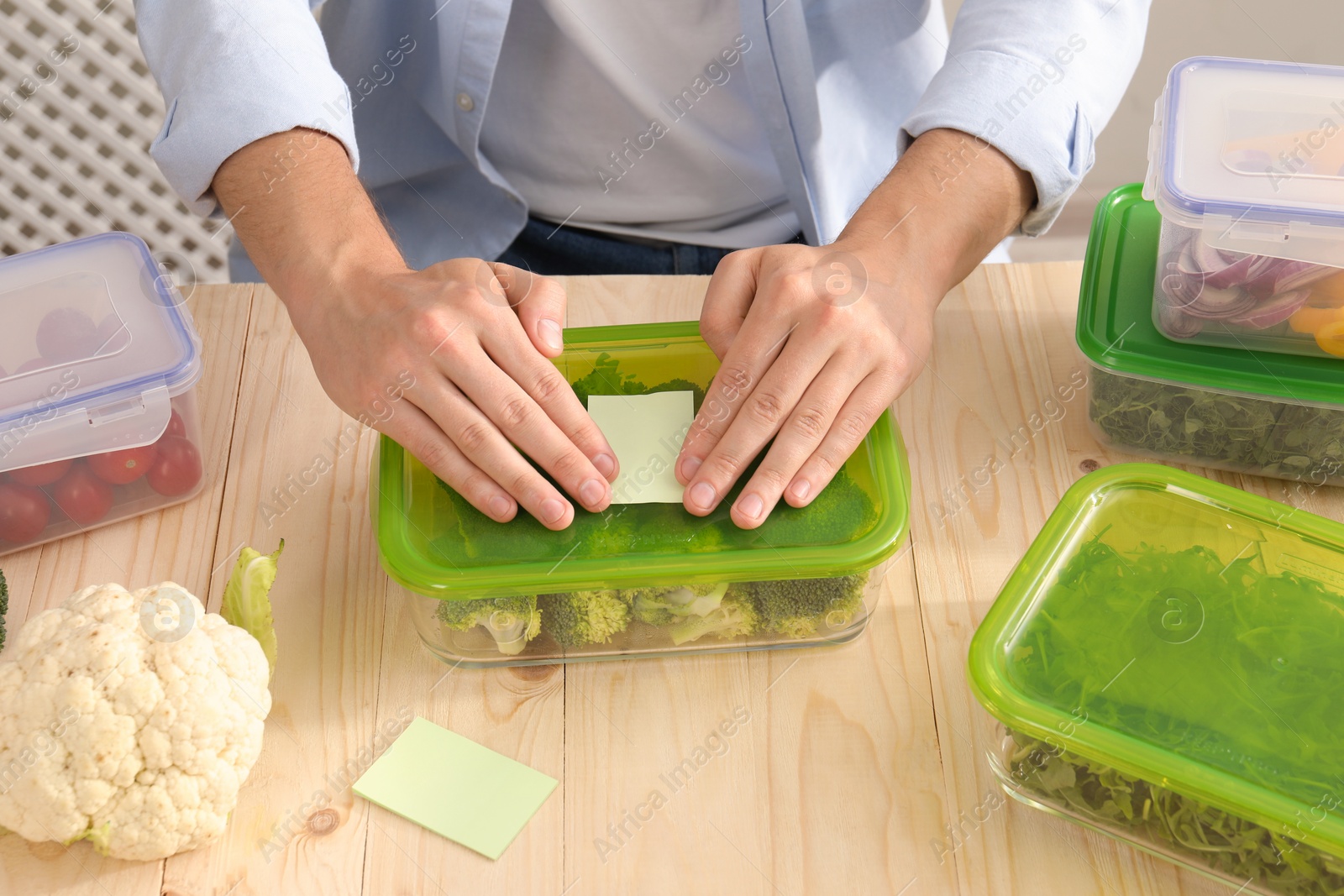 Photo of Man sticking paper note onto container with fresh broccoli at wooden table, closeup. Food storage