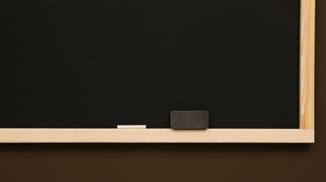 Photo of Clean blackboard with chalk and duster hanging on brown wall