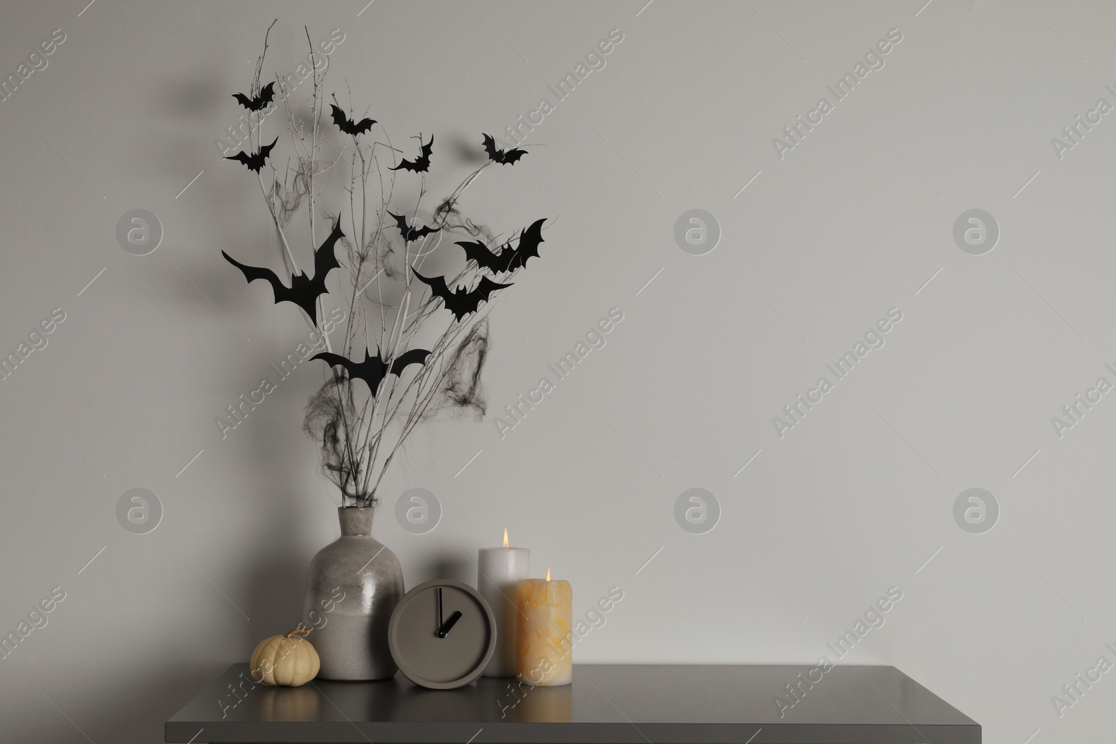 Photo of Alarm clock, burning candles and vase with paper bats  on table near white wall, space for text. Halloween decor