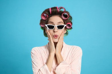 Beautiful young woman in bathrobe with hair curlers and sunglasses on light blue background