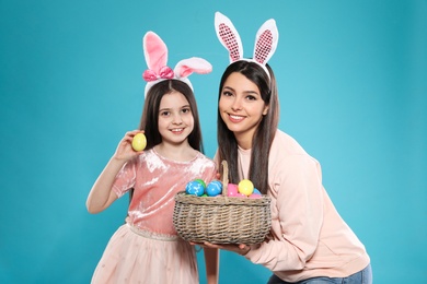 Mother and daughter in bunny ears headbands with basket of Easter eggs on color background