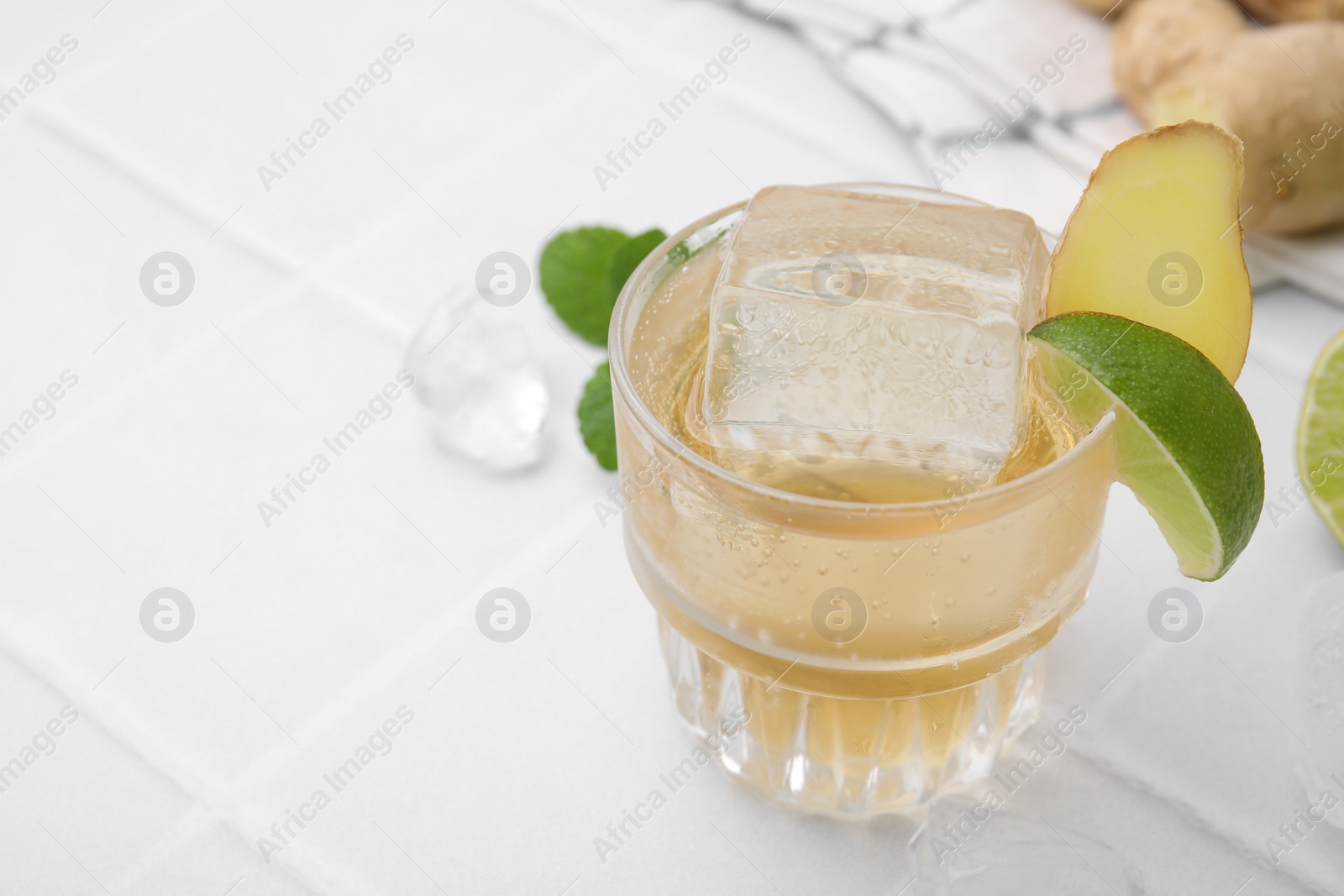 Photo of Glass of tasty ale with ice cube, lime and ginger slices on white tiled table, closeup. Space for text