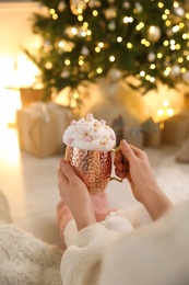 Photo of Woman holding cupdelicious drink with whipped cream and marshmallows near Christmas tree indoors, closeup