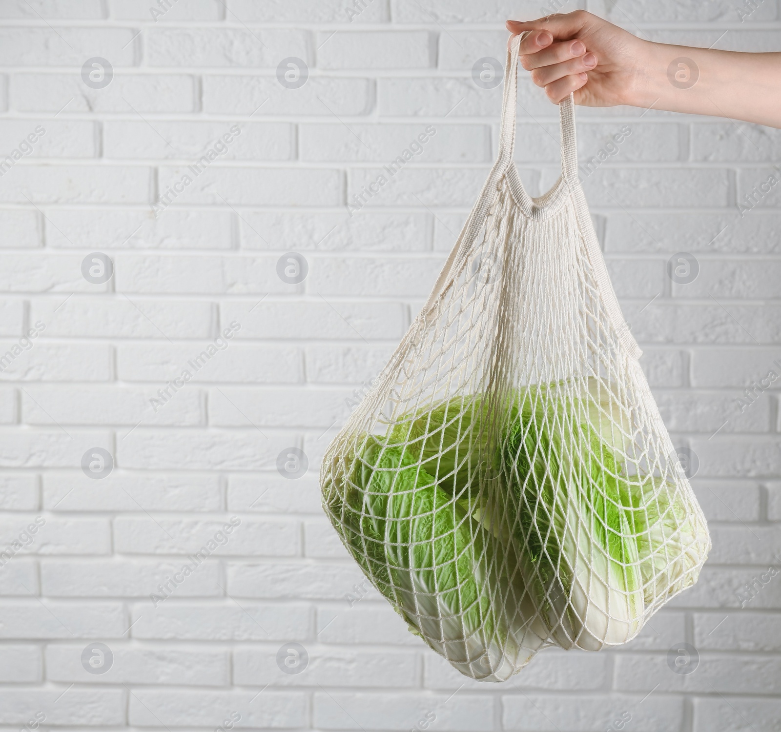 Photo of Woman holding string bag with fresh Chinese cabbages near white brick wall, closeup. Space for text