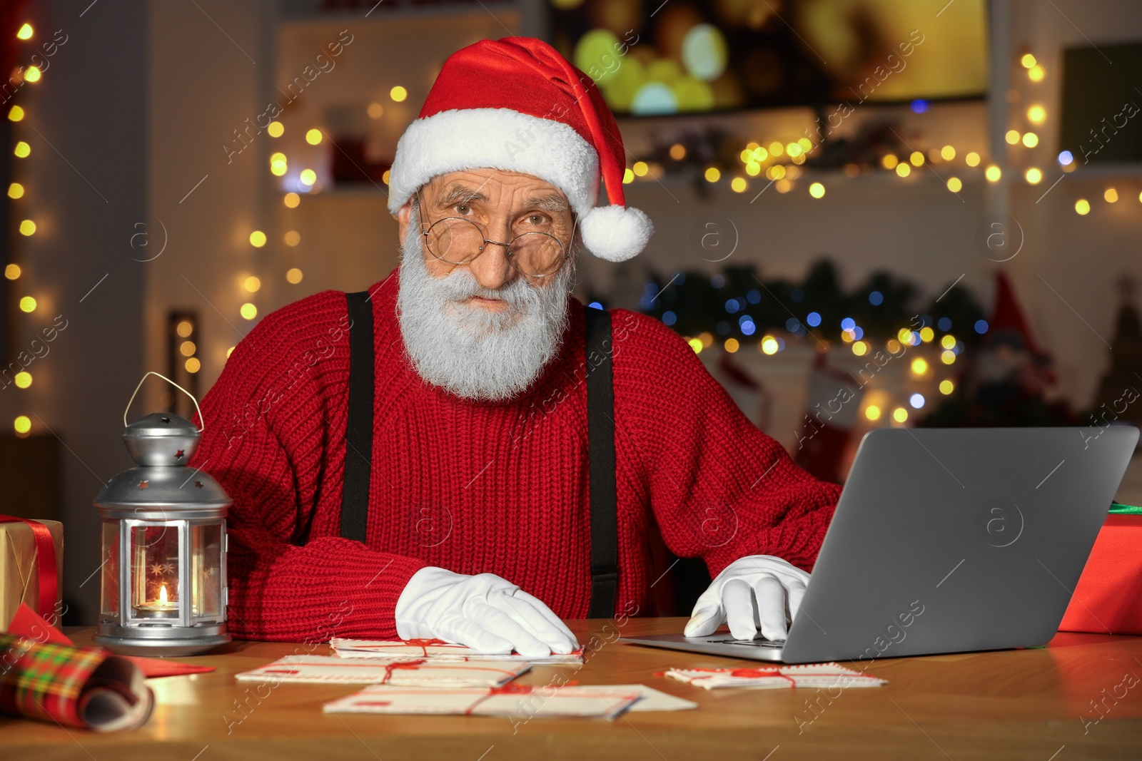 Photo of Santa Claus at his workplace. Letters and laptop on table in room decorated for Christmas