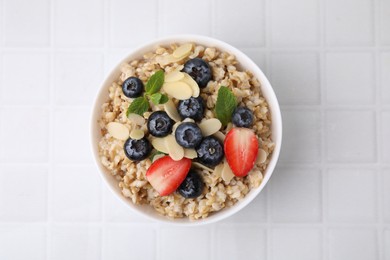 Photo of Tasty oatmeal with strawberries, blueberries and almond petals in bowl on white tiled table, top view