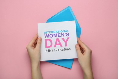 Image of Woman holding card with phrase International Women's Day and hashtag BreakTheBias on pink background, top view