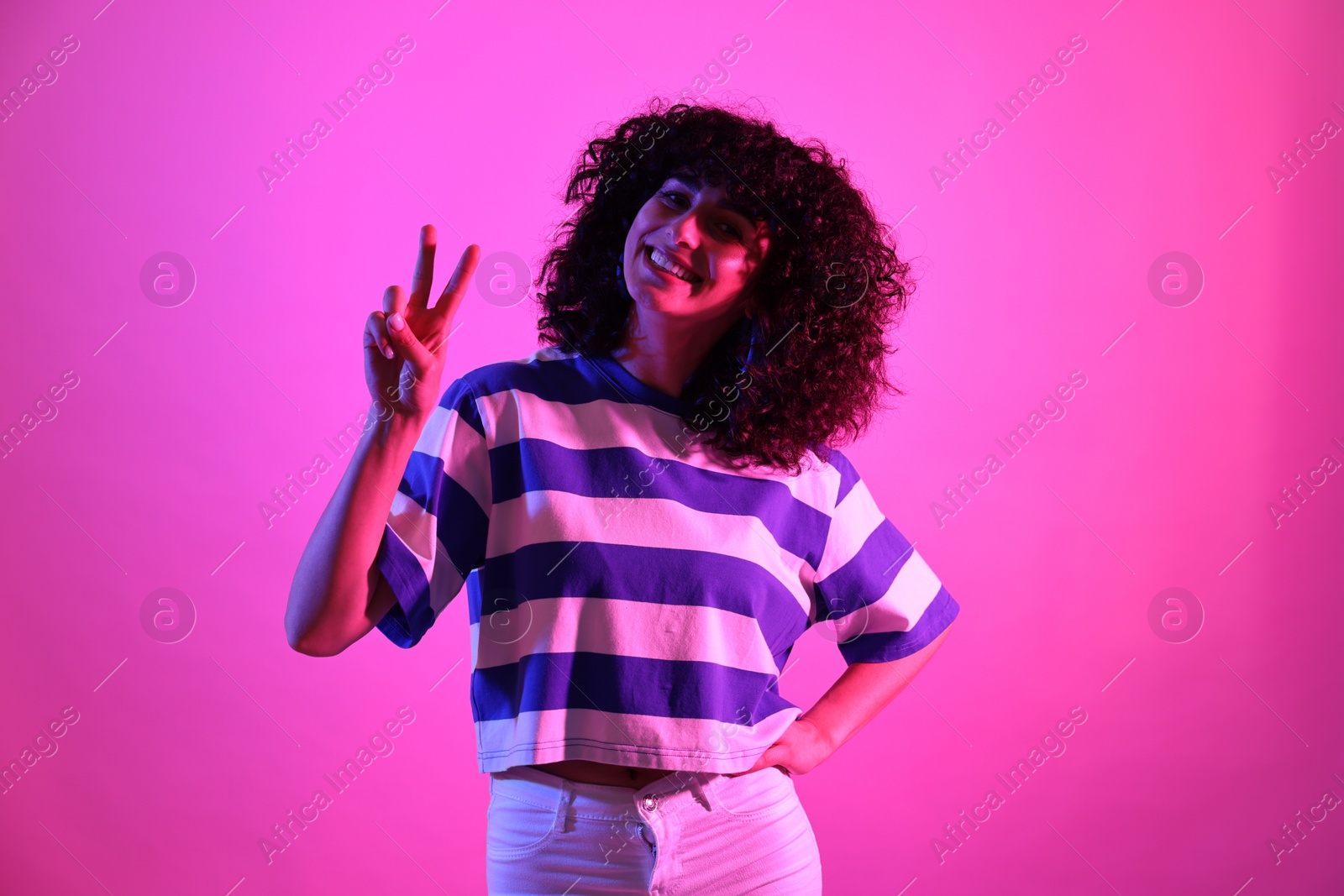 Photo of Beautiful young woman showing V-sign on color background in neon lights