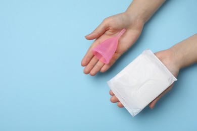 Photo of Woman holding menstrual cup and disposable pad on light blue background, top view. Space for text