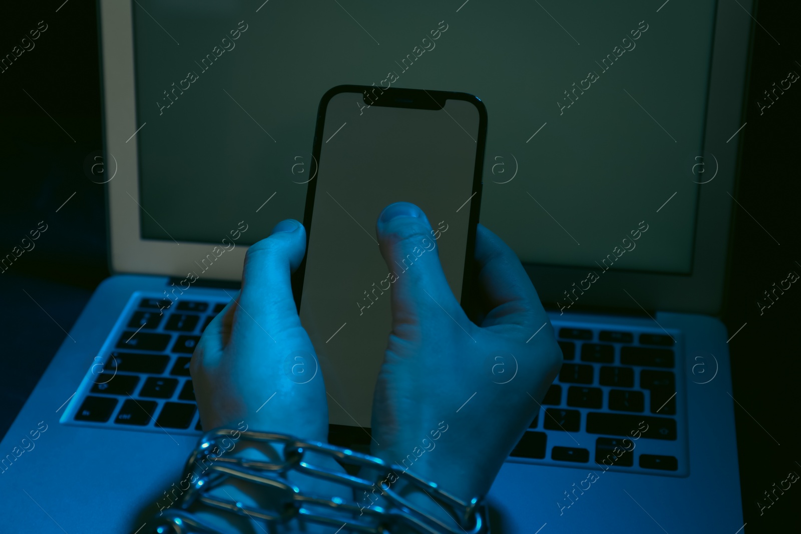 Photo of Woman with chained hands holding smartphone near laptop on dark background, closeup. Internet addiction