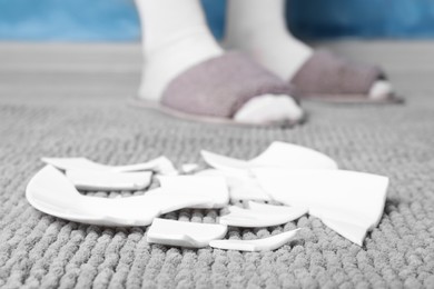 Photo of Woman in slippers standing near broken plate on rug at home, closeup