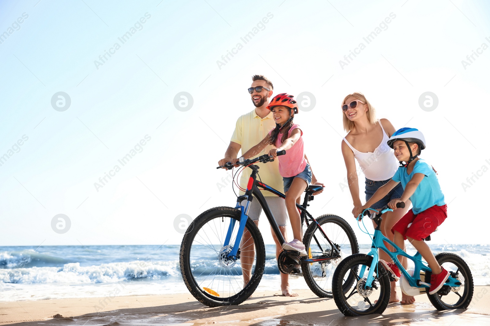 Photo of Happy parents teaching children to ride bicycles on sandy beach near sea