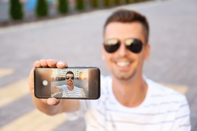 Photo of Young man taking selfie outdoors, focus on smartphone