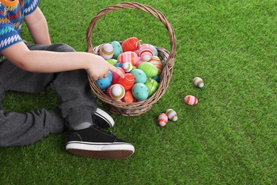 Photo of Little boy with basket full of dyed Easter eggs on green grass, closeup