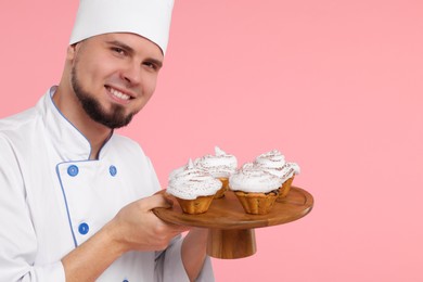 Happy professional confectioner in uniform holding delicious cupcakes on pink background