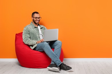 Photo of Smiling young man working with laptop on beanbag chair near orange wall, space for text