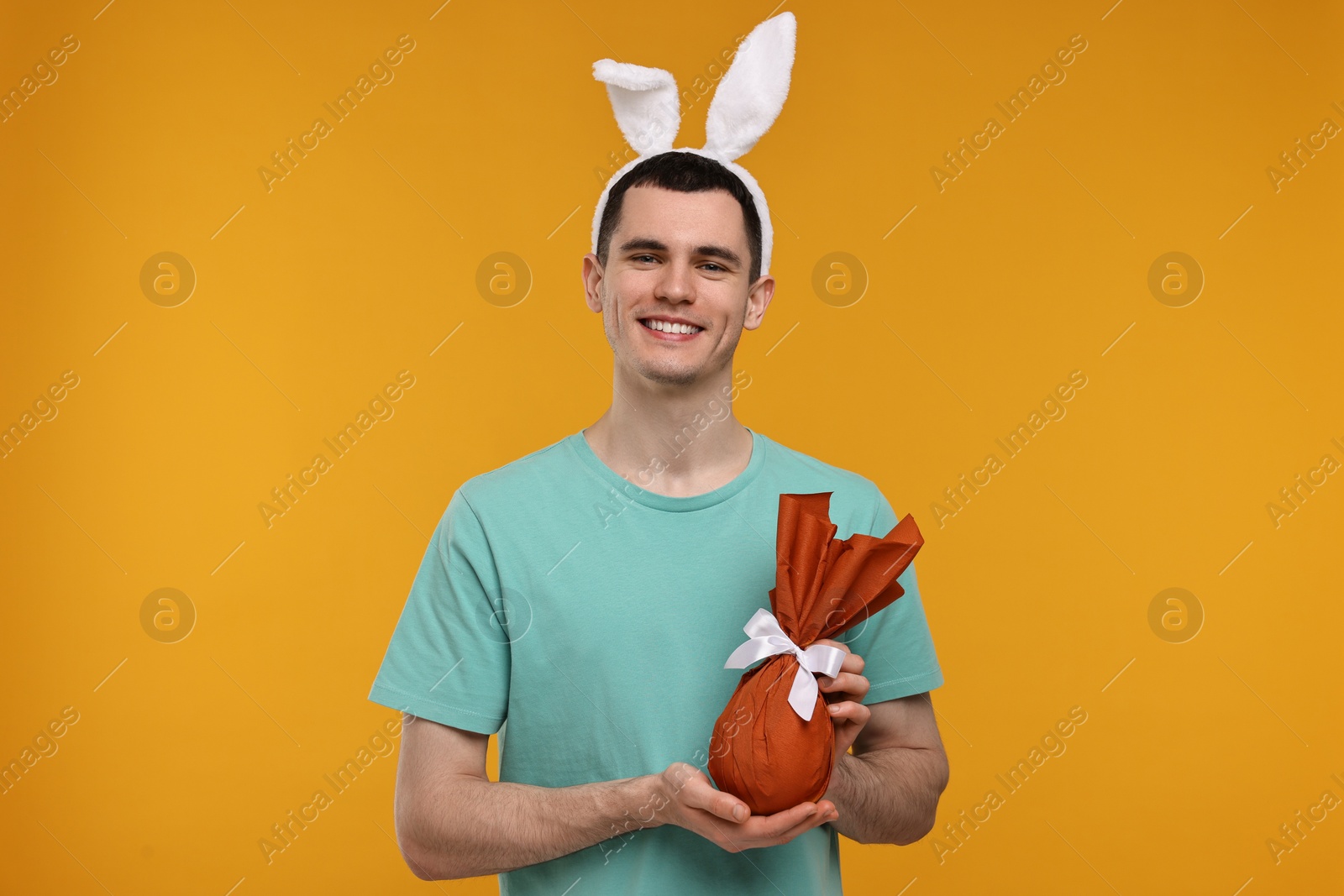 Photo of Easter celebration. Handsome young man with bunny ears holding wrapped gift on orange background