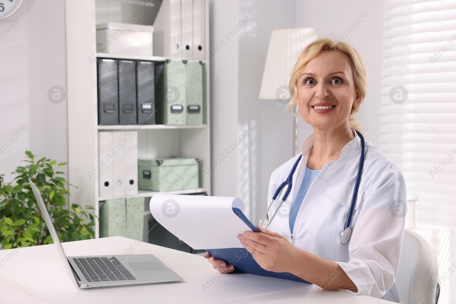 Photo of Doctor with laptop and clipboard at white table in clinic. Patient consultation