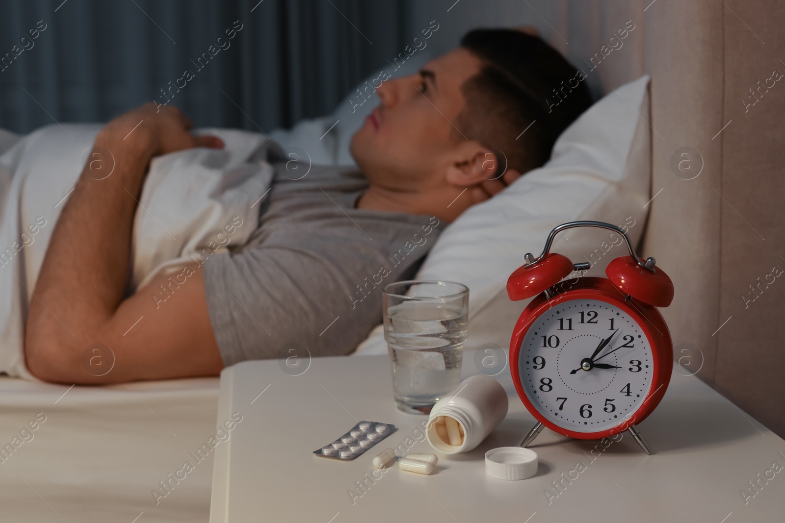 Photo of Man suffering from insomnia in bed at home, focus on pills and alarm clock