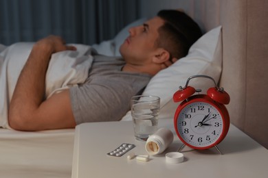 Photo of Man suffering from insomnia in bed at home, focus on pills and alarm clock