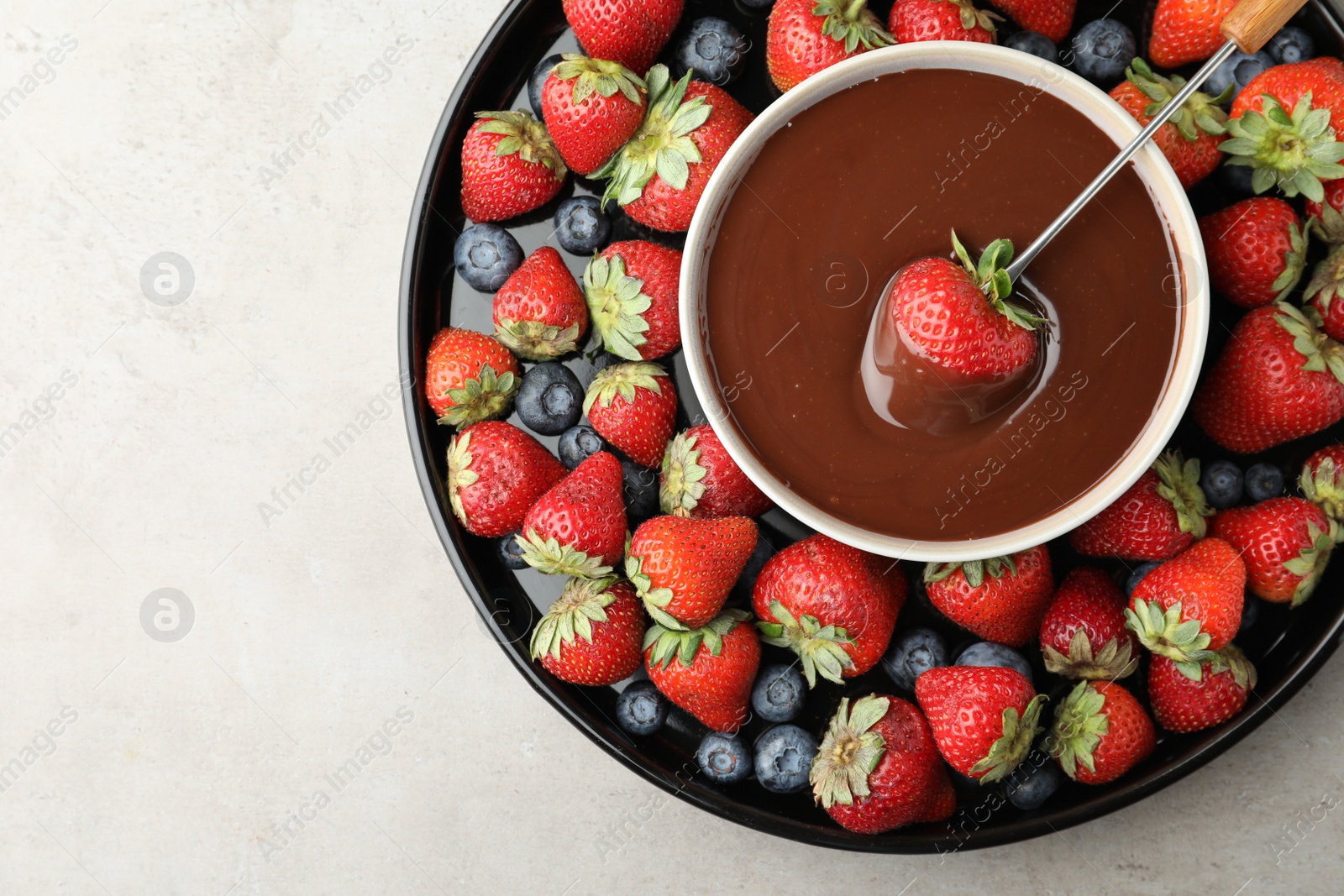 Photo of Fondue fork with strawberry in bowl of melted chocolate surrounded by different berries on light table, top view