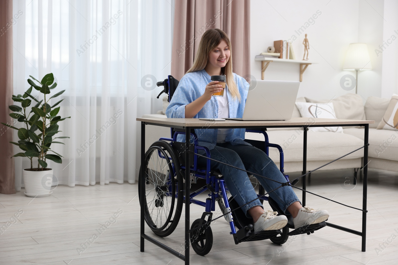 Photo of Woman in wheelchair with cup of drink using laptop at table in home office