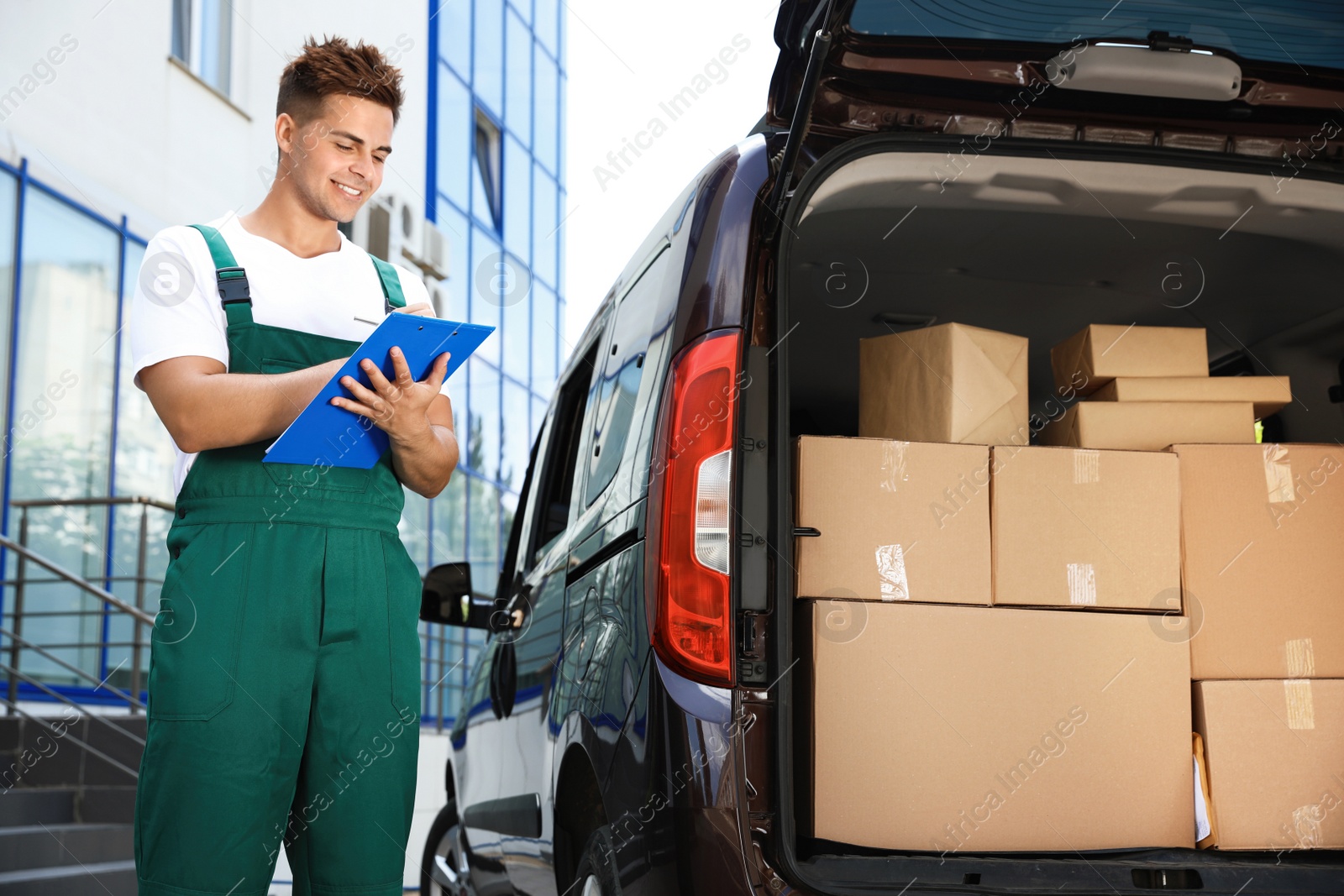 Photo of Young courier holding clipboard near delivery van with parcels outdoors