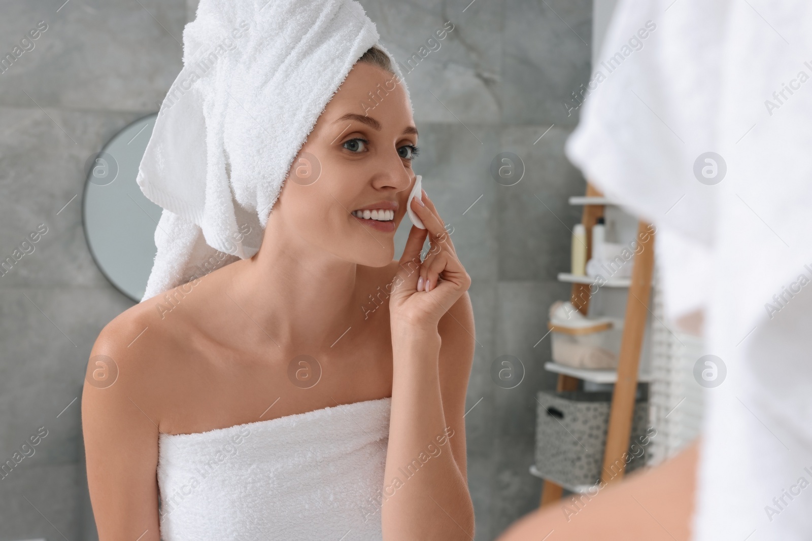 Photo of Young woman cleaning her face with cotton pad near mirror in bathroom