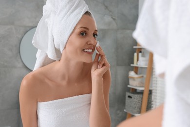 Photo of Young woman cleaning her face with cotton pad near mirror in bathroom