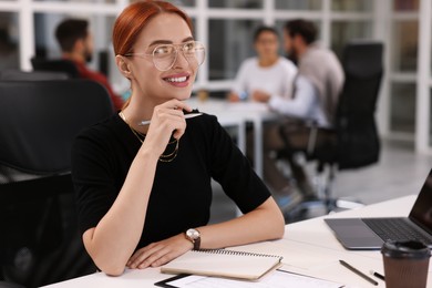 Photo of Team of employees working together in office. Happy woman with pen and notebook at table indoors