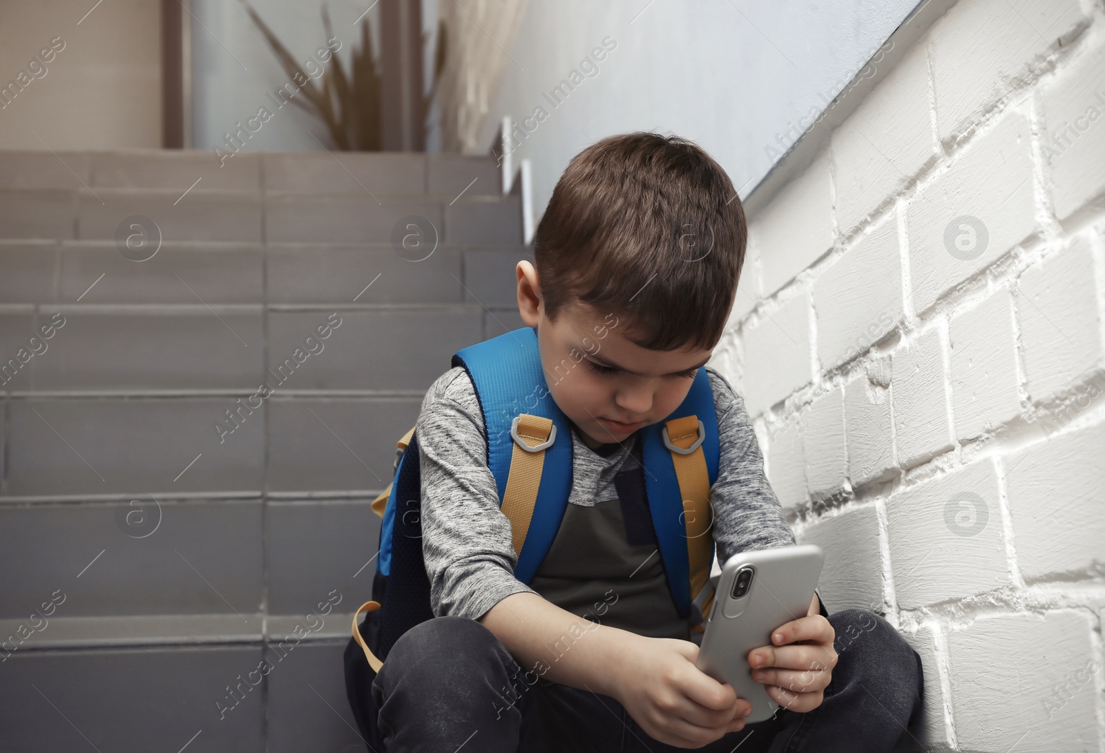 Photo of Sad little boy with mobile phone sitting on stairs indoors