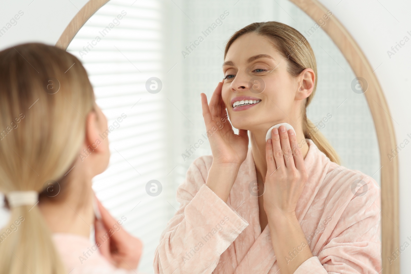 Photo of Beautiful woman removing makeup with cotton pad near mirror indoors