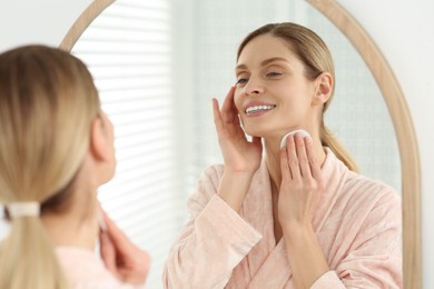 Photo of Beautiful woman removing makeup with cotton pad near mirror indoors