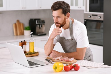 Photo of Man making dinner while watching online cooking course via laptop in kitchen
