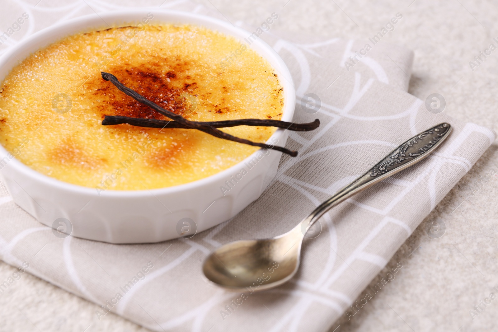 Photo of Delicious creme brulee in bowl, vanilla pods and spoon on light textured table, closeup