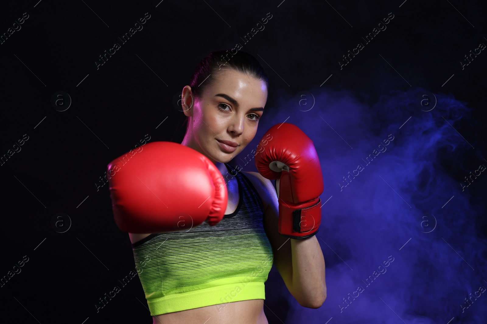 Photo of Portrait of beautiful woman wearing boxing gloves in color lights and smoke on black background