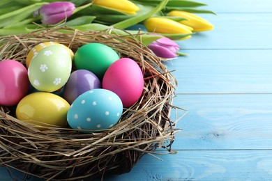 Photo of Colorful eggs in nest and tulips on blue wooden background, closeup. Happy Easter
