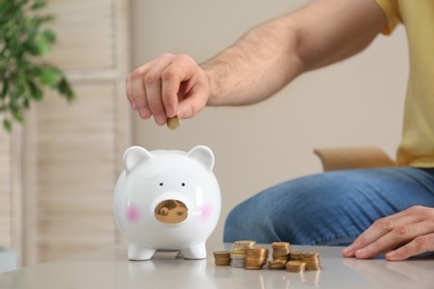 Man putting coin into piggy bank at table indoors, closeup. Money saving