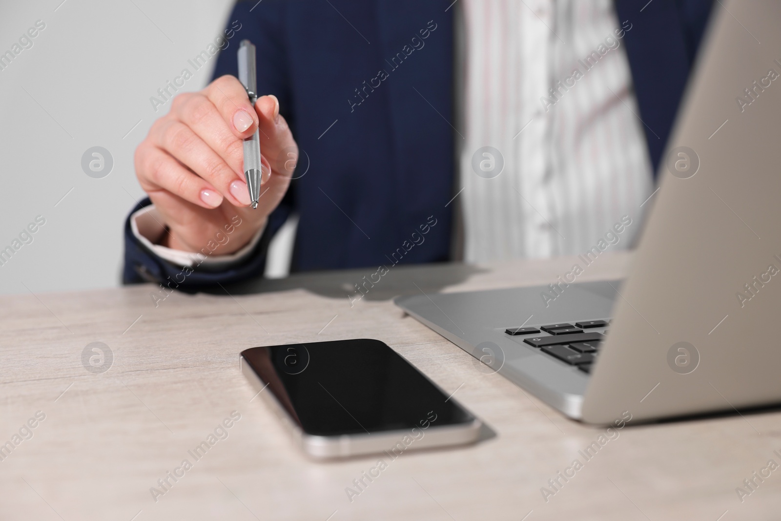 Photo of Woman with smartphone and pen working on laptop at wooden table, closeup. Electronic document management