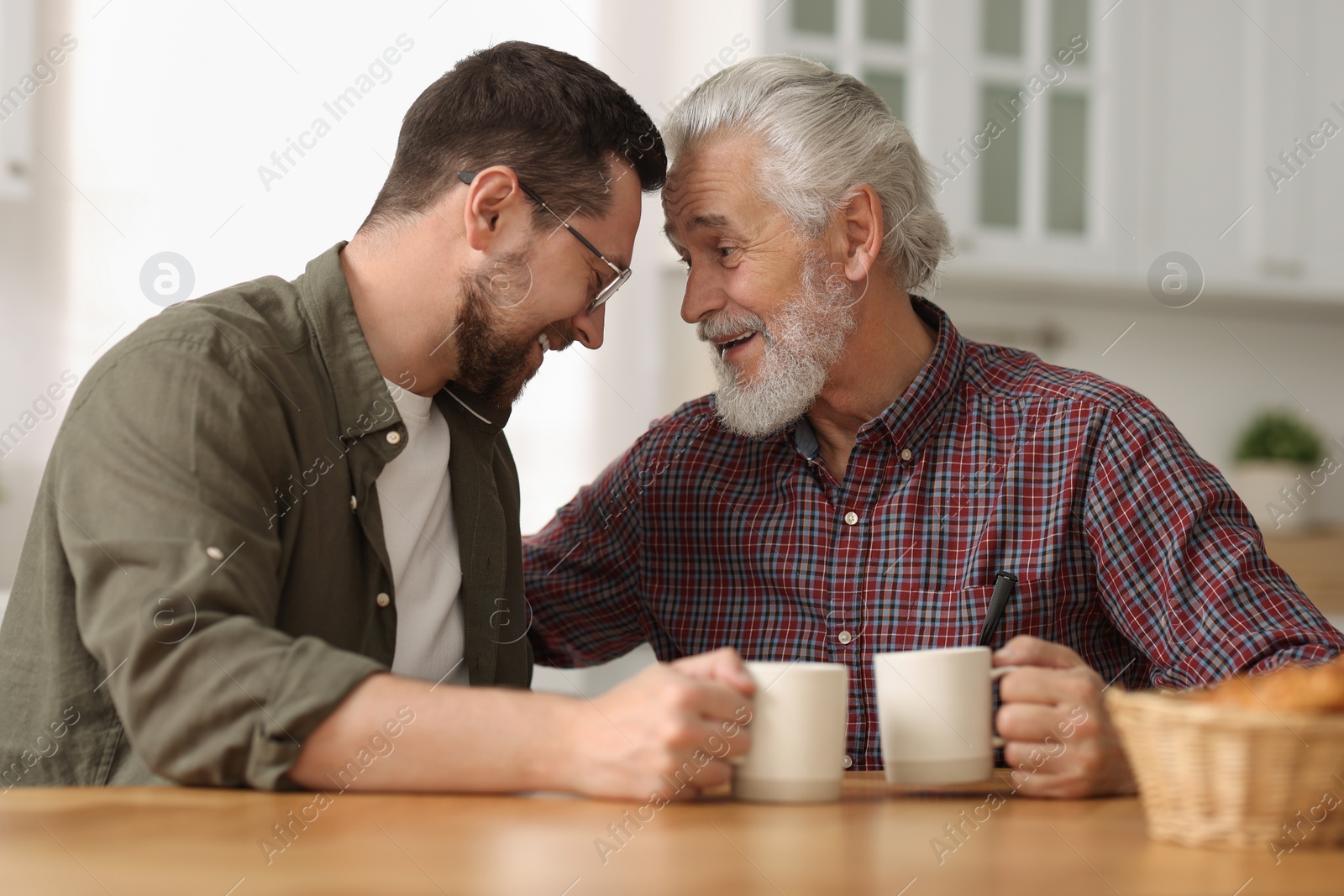 Photo of Happy son and his dad with cups at wooden table in kitchen