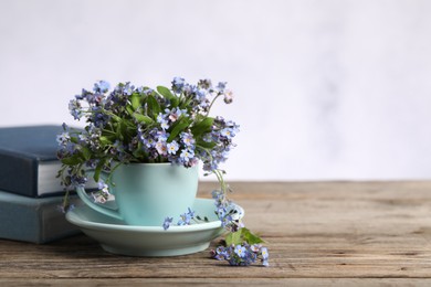 Photo of Beautiful forget-me-not flowers in cup, saucer and books on wooden table against light background, closeup. Space for text