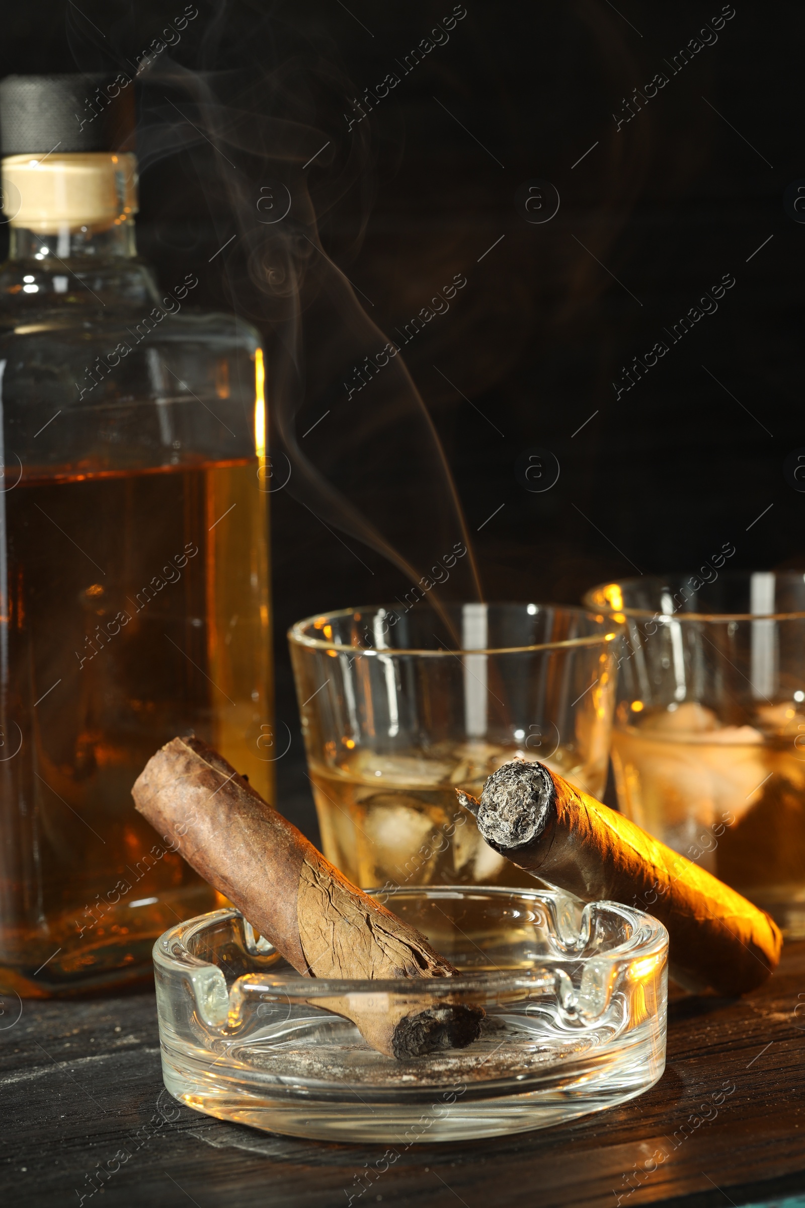 Photo of Cigars, ashtray and whiskey with ice cubes on black wooden table