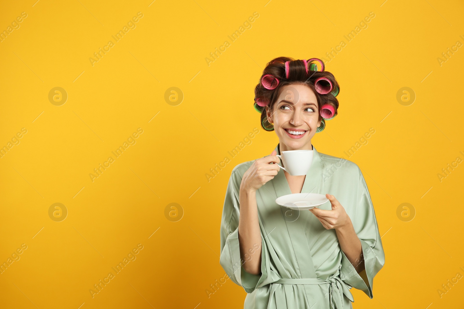 Photo of Beautiful young woman in silk bathrobe with hair curlers and cup of drink on yellow background. Space for text